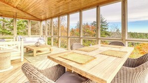 Uma área de estar ao ar livre coberta, com uma mesa e cadeiras de madeira, janelas amplas e vista para árvores e céu.