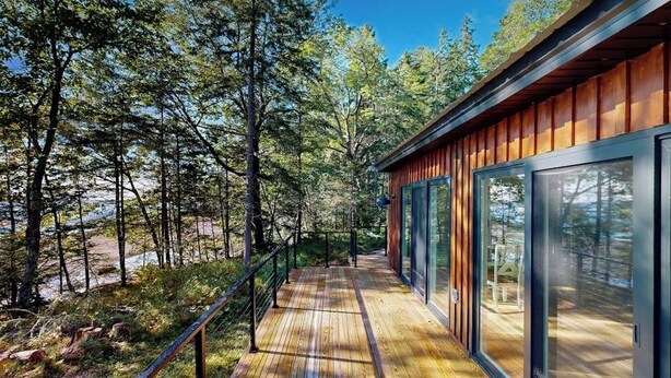A wooden deck with glass doors leading to a modern building surrounded by trees. - Modern Maine Cottage With River Views, Deck, and a Convenient Washer/dryer (Stockton Springs)
