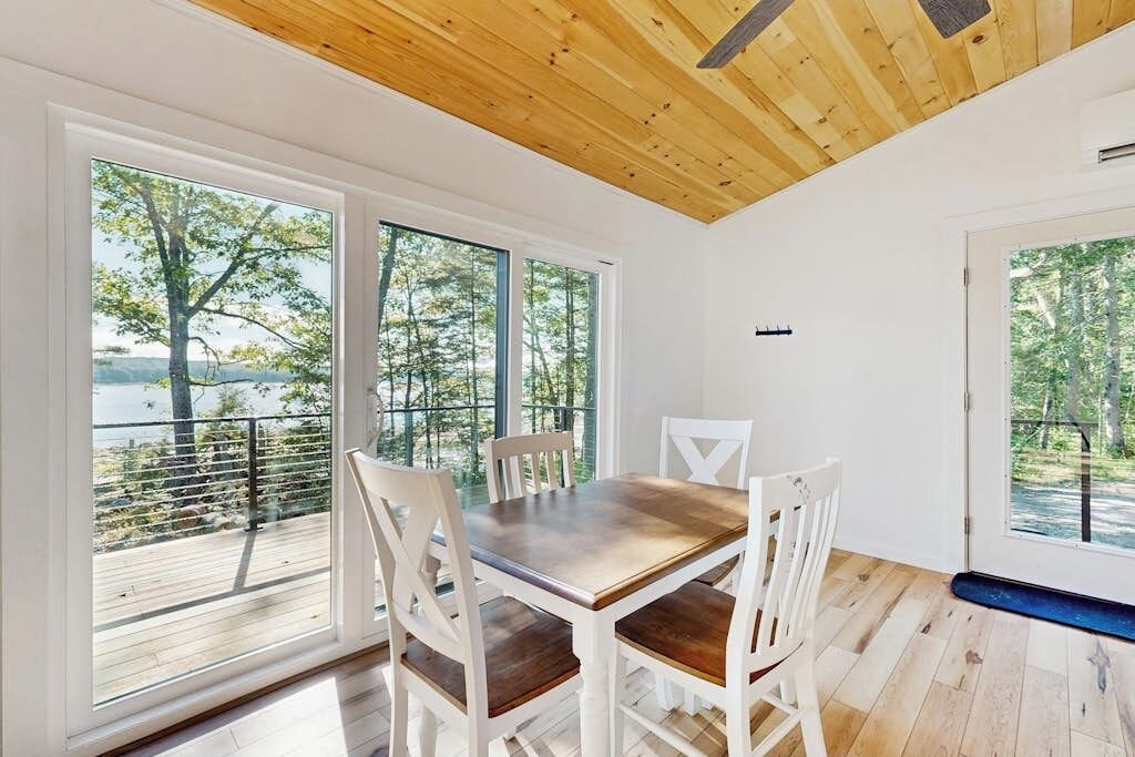 Un'area pranzo con tavolo in legno e sedie bianche, ampie finestre vista alberi e uno specchio d'acqua, e soffitto in legno.