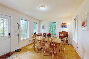 A dining room with a wooden table and chairs, a coat rack, and a bookshelf with books and decorative items. - Cozy Apartment With Harbor View - Full Kitchen & Washer/dryer - Walk to Water (Rockport)