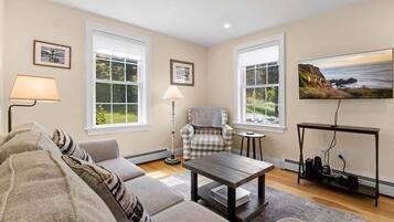 A living room with a sofa, armchair, coffee table, lamp, TV, and large windows.