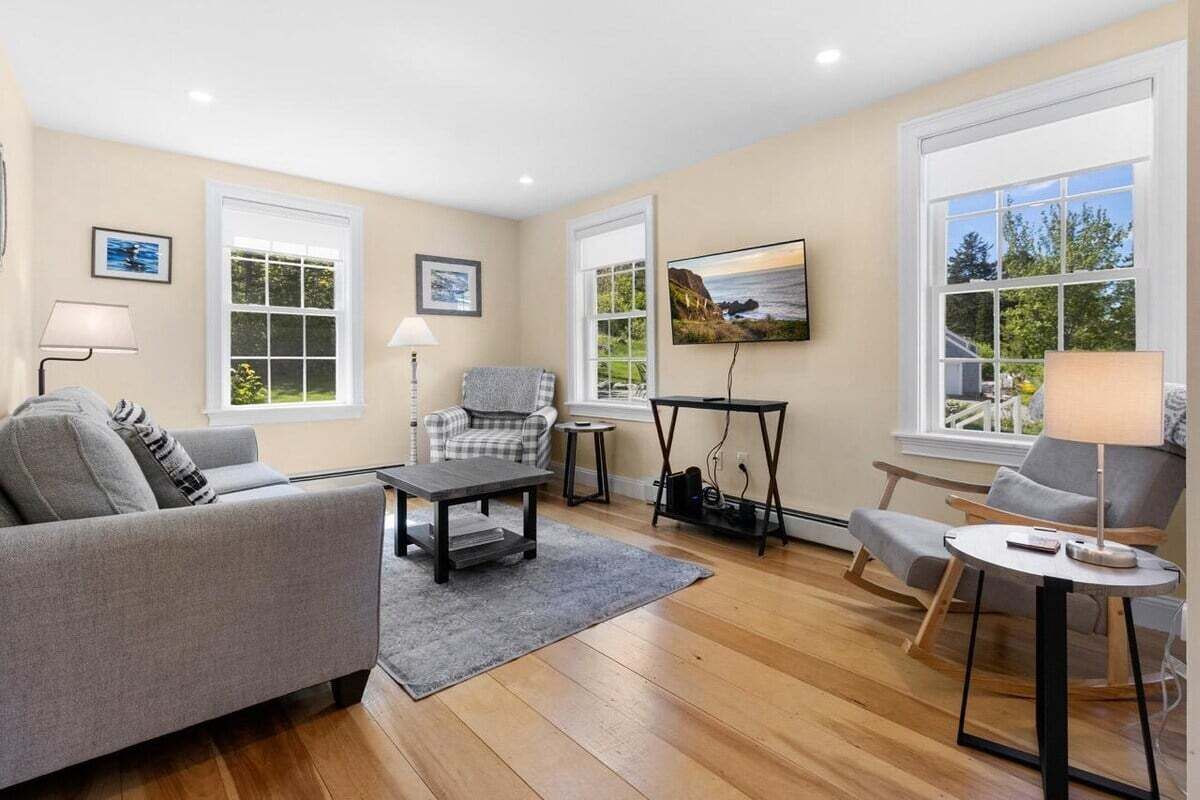 A living room with a sofa, a coffee table, a chair, a small table, and a TV. There are windows with curtains, a lamp, and a picture on the wall.