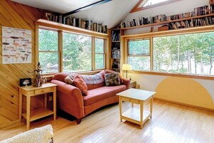 Um quarto aconchegante com um sofá vermelho, uma mesinha, uma estante de livros e grandes janelas com vista para as árvores.
