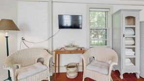 A room with two wicker chairs, a small wooden table, a television mounted on the wall, and a white cabinet with shelves. - Downtown, Ground Floor Studio Near the Ocean W/patio, Outdoor Dining Area & More (Wiscasset)