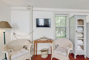 A room with two wicker chairs, a small wooden table, a television mounted on the wall, and a white cabinet with shelves.