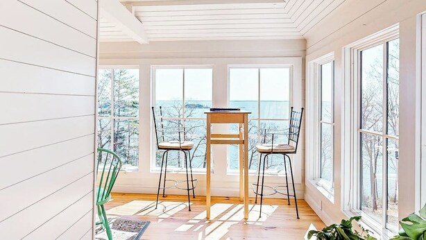 Ein Zimmer mit Holzboden, einem Tisch mit Stühlen, großen Fenstern und Blick auf Bäume.
