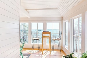 Una habitación con piso de madera, una mesa con sillas, ventanas grandes y vista a árboles.