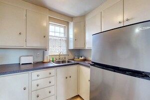 A kitchen with white cabinets, a stainless steel refrigerator, a sink, and a window with lace curtains.