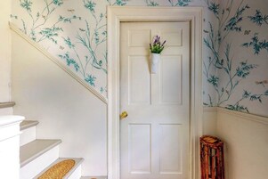 A white door with a small plant hanging on it, a wooden stool to the right, and a staircase with a woven mat on the bottom step.