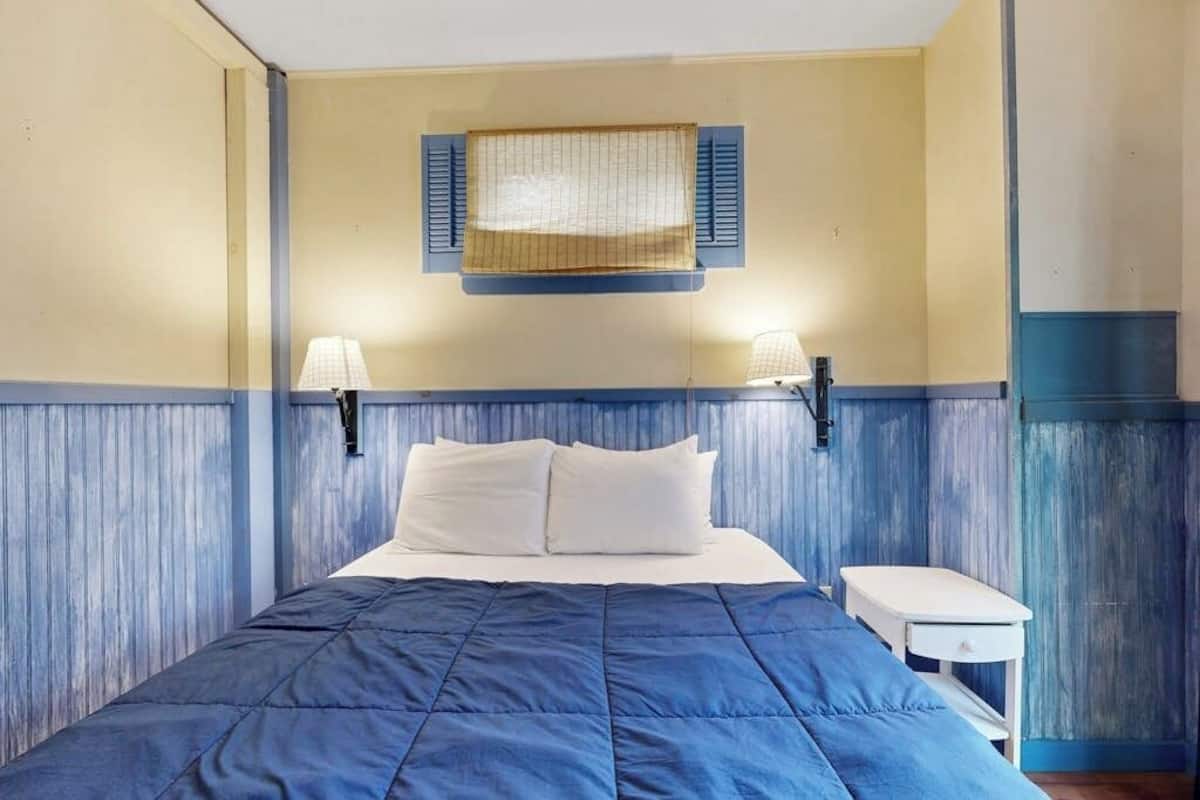 A bedroom with a bed, two bedside lamps, a small table, and a window with shutters.