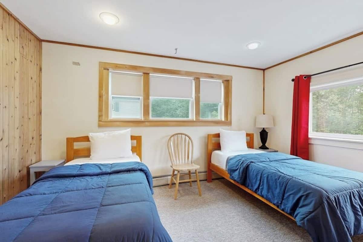 A room with two beds, a window with blinds, a chair, and a nightstand.