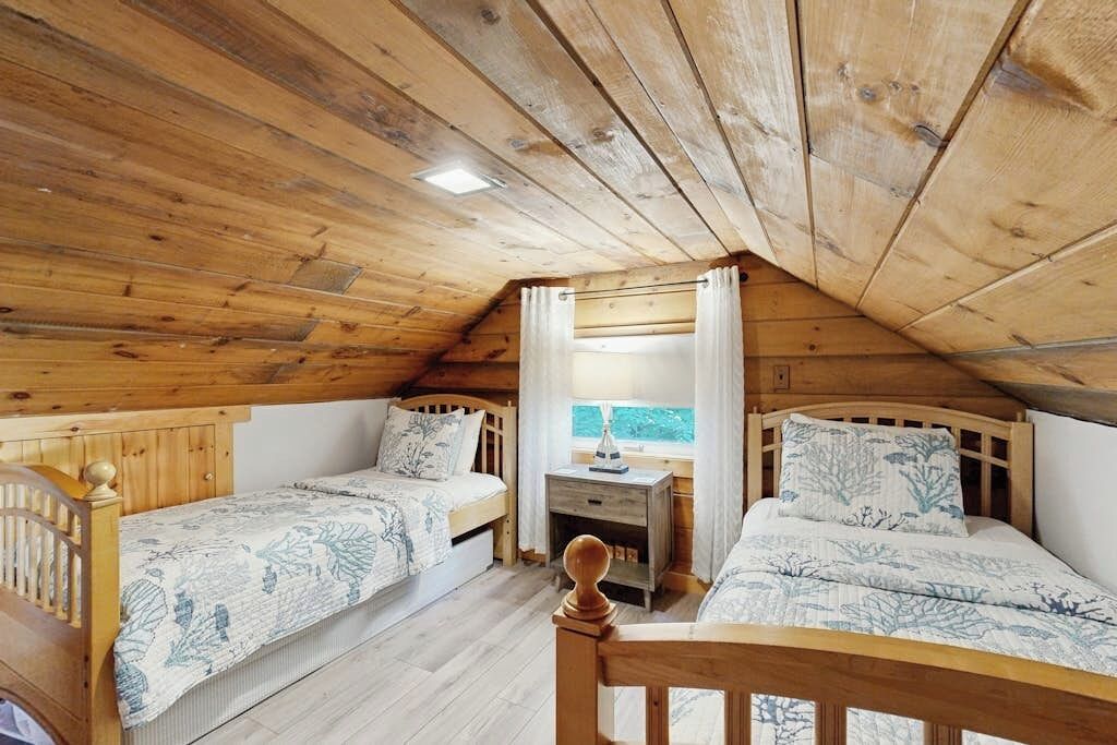 A cozy attic bedroom with two wooden beds, a small desk, and a window with curtains.