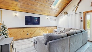 A cozy living room with a wooden ceiling, a grey sofa, a flat-screen TV, and a small Christmas tree. - Scenic Retreat With a Spacious Deck, gas Fireplace, and Moosehead Lake Views (Greenville)