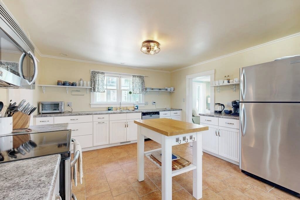 A kitchen with white cabinets, a stainless steel refrigerator, a wooden table, and a window with curtains.