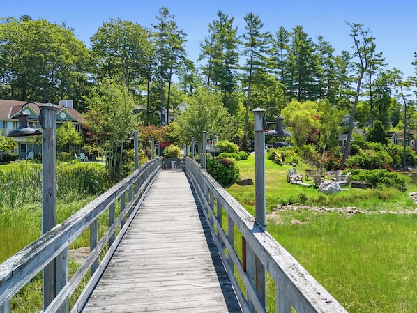A wooden bridge leading to a residential area with houses, a pond, and greenery. - Mabel's Maine Studio on the River (Edgecomb)