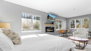 Ein Schlafzimmer mit einem Bett, einem an der Wand montierten Fernseher, einem Kamin, einer Essecke mit Tisch und Stühlen sowie großen Fenstern.