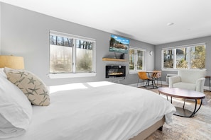A bedroom with a bed, a TV mounted on the wall, a fireplace, a dining area with a table and chairs, and large windows. - Mabel's Maine Studio on the River (Edgecomb)