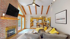 A modern living room with a fireplace, a flat-screen TV, a ceiling fan, and a dining area visible through an open door. - Woodland Home With Screened Porch, 2 Living Rooms - Near Historic Waterfront (Bath)