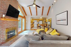 A modern living room with a fireplace, a flat-screen TV, a ceiling fan, and a dining area visible through an open door. - Woodland Home With Screened Porch, 2 Living Rooms - Near Historic Waterfront (Bath)