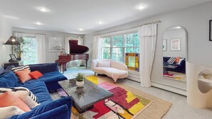 A living room with a blue sofa, a coffee table, a piano, and a large window with curtains. - Woodland Home With Screened Porch, 2 Living Rooms - Near Historic Waterfront (Bath)