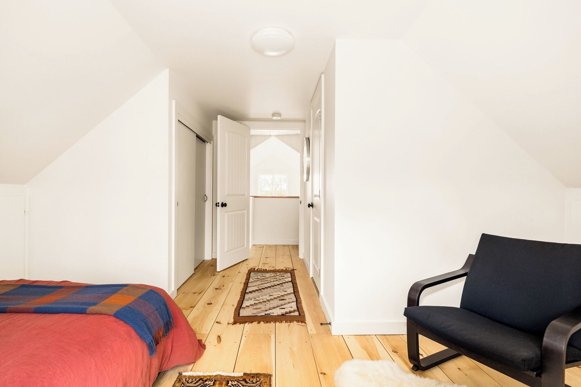 A bedroom with a bed, a chair, a wooden floor, and a hallway leading to a window.