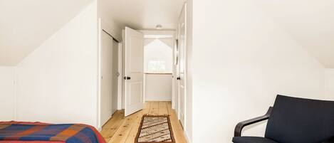 A bedroom with a bed, a chair, a wooden floor, and a hallway leading to a window.