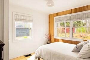 Un dormitorio con cama, ventana con persianas, una mesita con un florero y un panel de pared de madera.