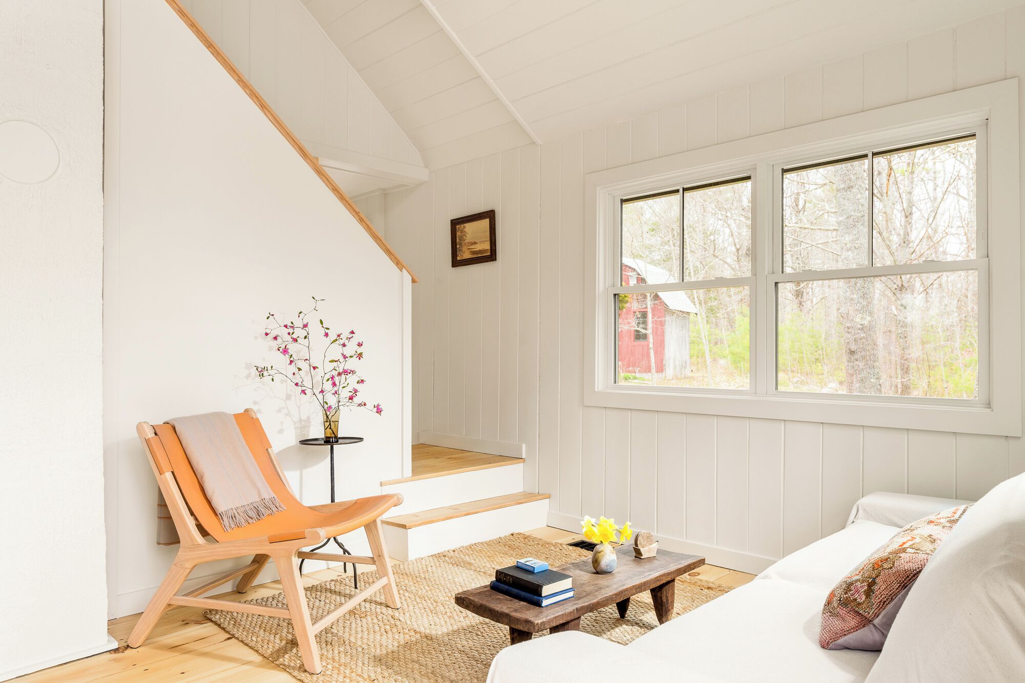 A room with a white wall, a wooden chair, a sofa, a coffee table, and a window with a view of trees.