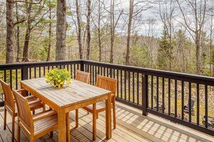 Una terraza de madera con una mesa y sillas, rodeada por una barandilla negra, con vistas a una zona boscosa.