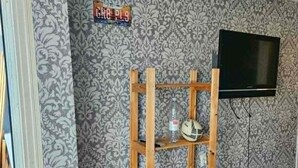 A room with patterned wallpaper, a wooden shelf unit, a television mounted on the wall, and a small stool. - Beautiful 1-bedroom apartment in charming Liepāja perfect for relaxing stay (Liepāja)