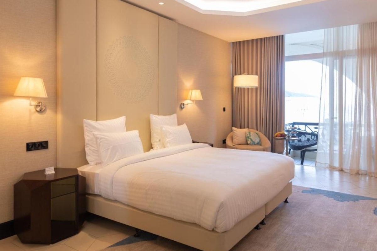 Una habitación de hotel con una cama grande, dos lámparas de noche, una silla, una mesita y vistas al exterior a través de una ventana con cortinas.