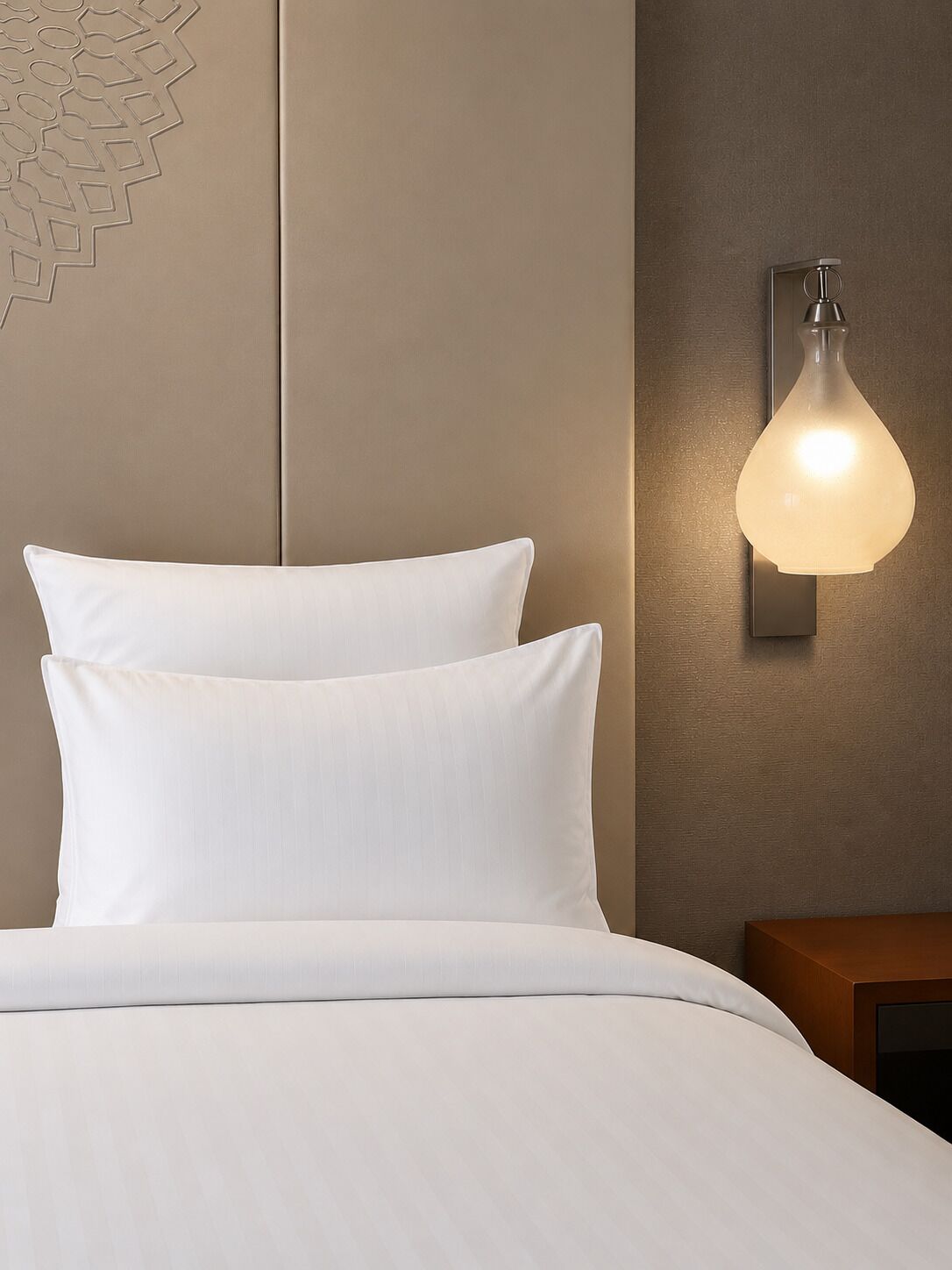 Uma cama com roupa de cama branca, uma luminária na parede e uma mesinha de cabeceira de madeira.