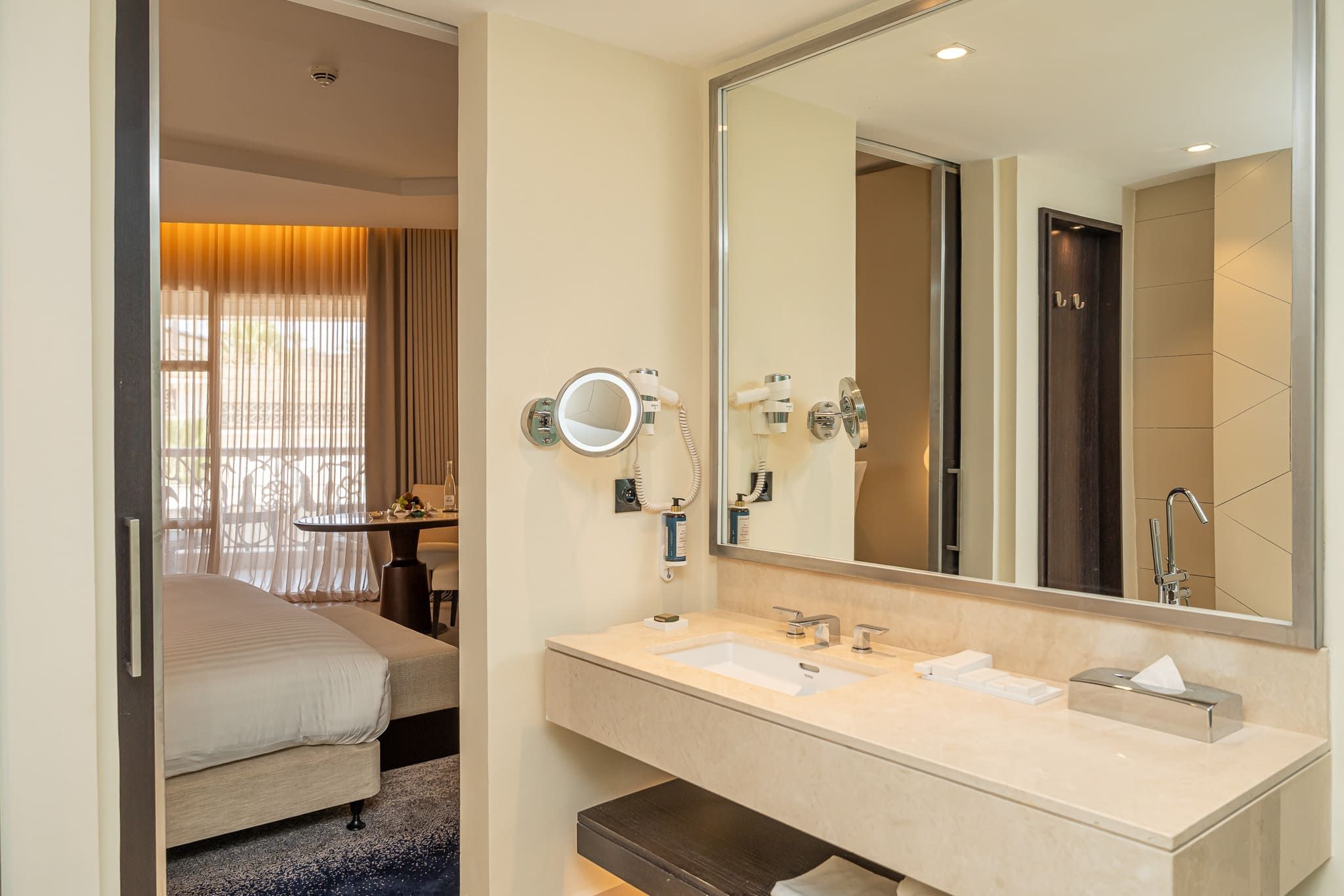 Habitación de hotel con cama, baño con lavamanos doble, espejo y secador de pelo.