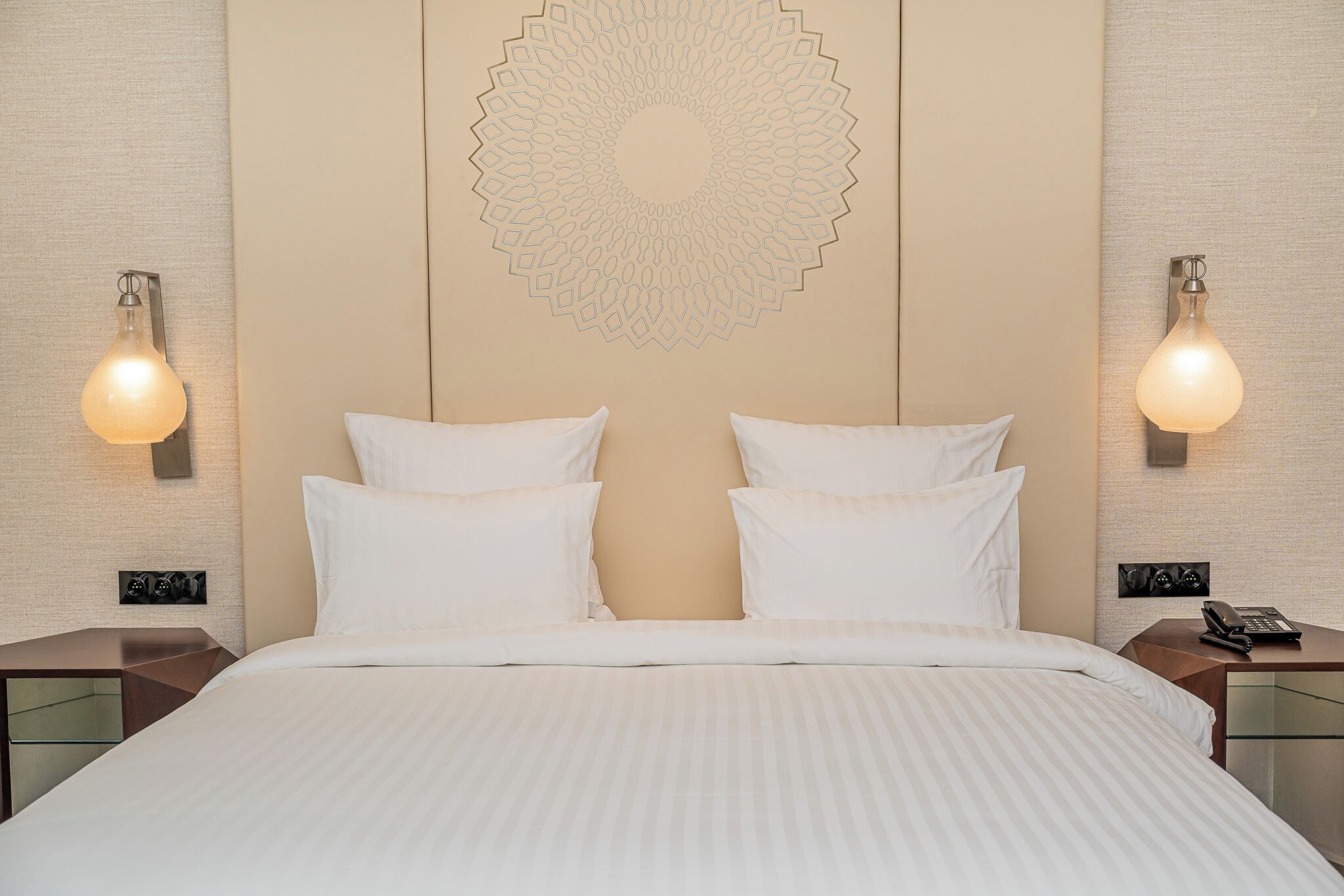 A hotel room with a large bed, two bedside tables, a wall-mounted decorative pattern, and wall sconces.