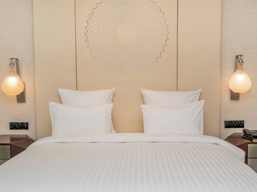 A hotel room with a large bed, two bedside tables, a wall-mounted decorative pattern, and wall sconces.