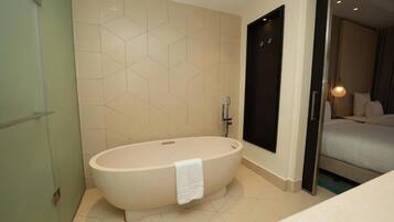 Un baño moderno con una bañera independiente, una cabina de ducha con paredes de vidrio y una cama visible en la habitación contigua.