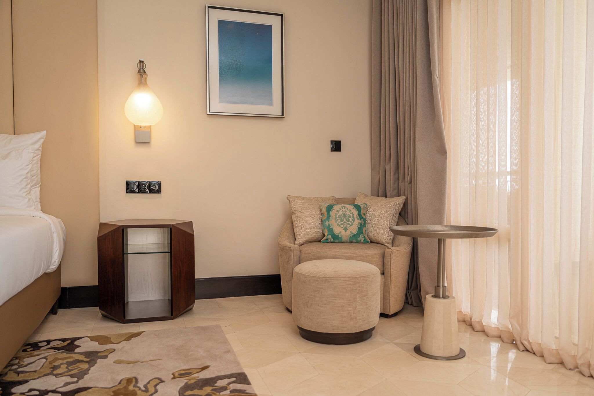 Habitación de hotel con cama, zona de estar con sofá y reposapiés, una mesita y una lámpara de pared, además de un cuadro enmarcado.