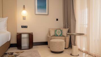 Habitación de hotel con cama, zona de estar con sofá y reposapiés, una mesita y una lámpara de pared, además de un cuadro enmarcado.