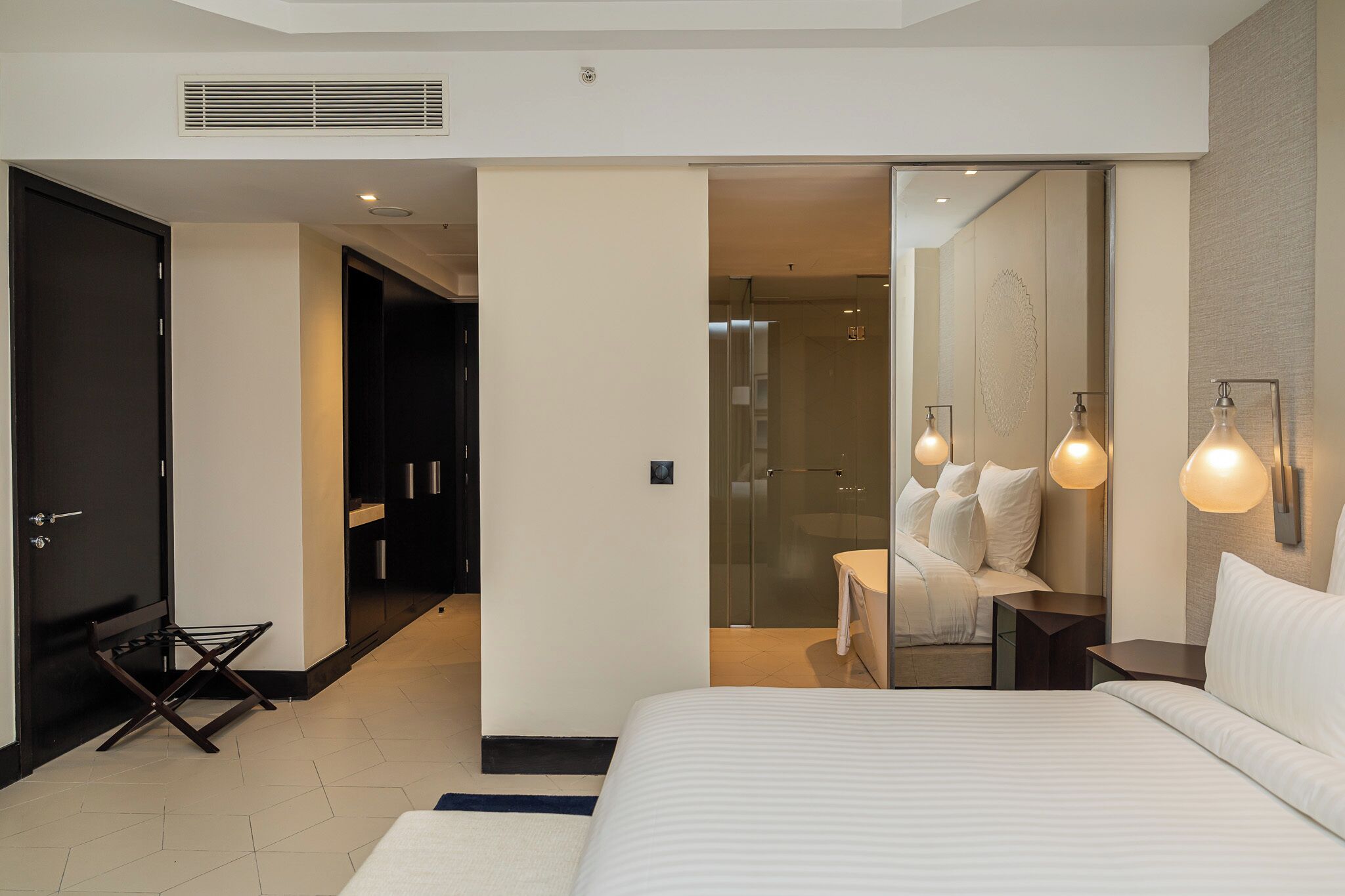 A modern hotel room with a large bed, a walk-in closet, a bathroom with a glass shower, and a seating area with a sofa and lamps.