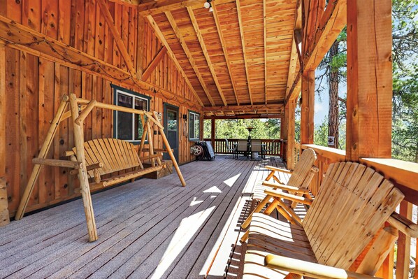 A wooden cabin with a porch, Adirondack chairs, a swing set, and a view of trees.