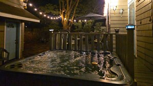 Un bain à remous avec une terrasse en bois et des guirlandes lumineuses, entouré d’arbres et d’une maison.