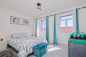 Ein Schlafzimmer mit Bett, Nachttisch, Nachttischlampe, einem Fenster mit Vorhängen und einem grünen Reisebett.