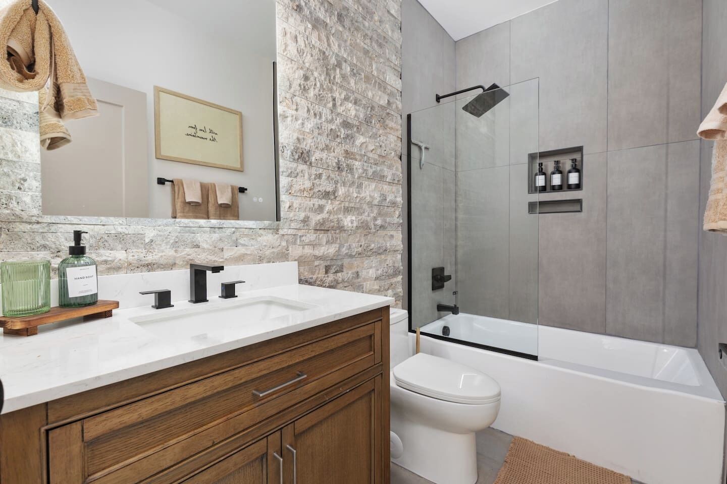 Un baño con pared de piedra, un espejo grande, lavamanos doble, inodoro y ducha con cerramiento de vidrio.