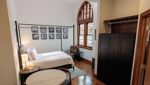 Un dormitorio con cama, mesita de noche, una silla, una mesa redonda, armario y una ventana con vidrieras, además de cuadros en la pared.