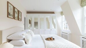 Un dormitorio con cama, almohadas, mesita de noche con lámpara, bandeja con aperitivos, armario y ventana con vistas.