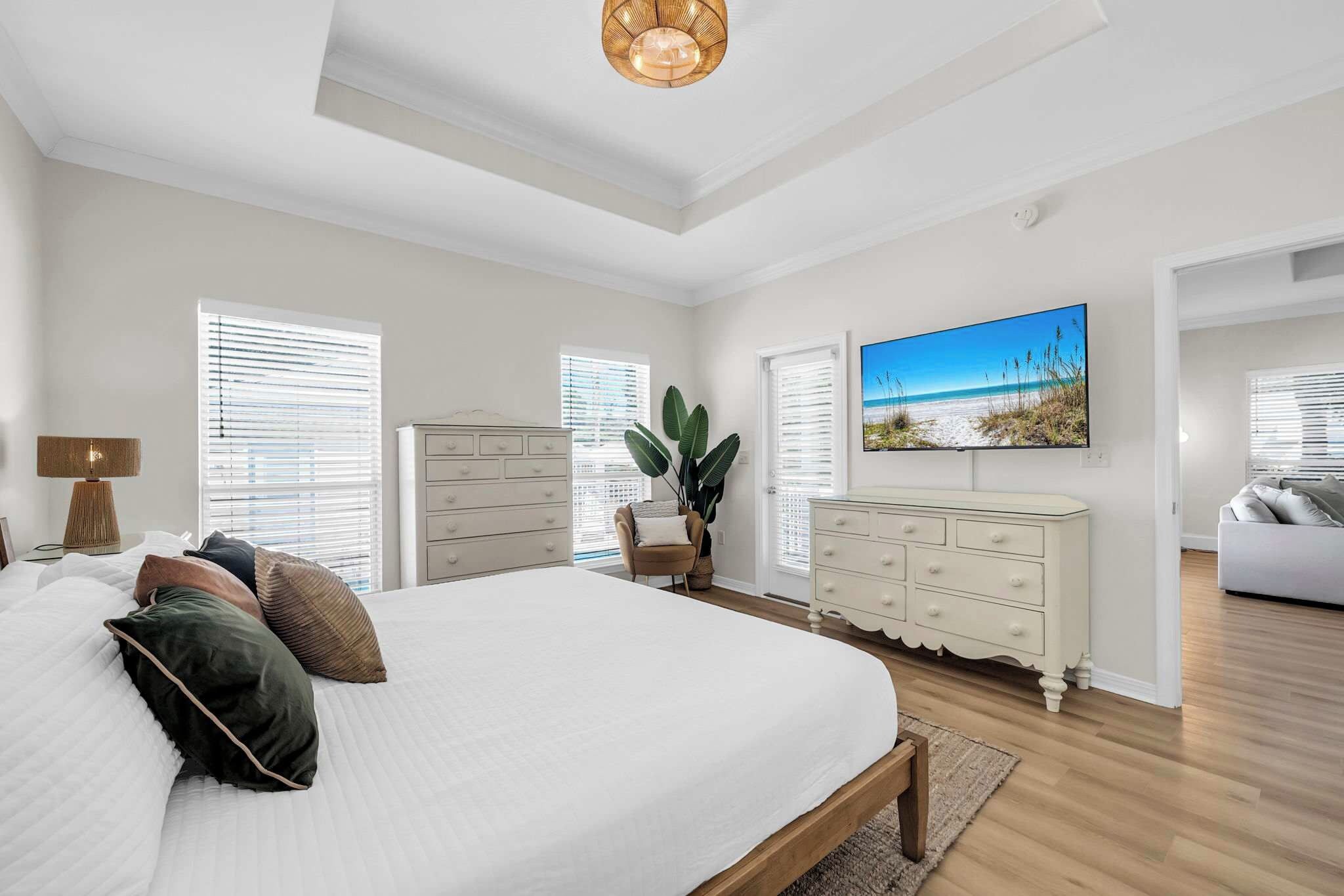 Une chambre à coucher avec un grand lit, une commode, une chaise, une plante et un tableau représentant une scène de plage.