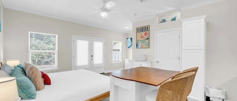 A bedroom with a bed, a desk, and a chair. There are two doors, a window, and a ceiling fan.