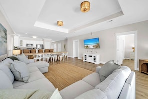 Ein modernes Wohnzimmer mit einer Eckcouch, einem Essbereich mit Tisch und Stühlen, einer Küche im Hintergrund und einem an der Wand befestigten Fernseher.