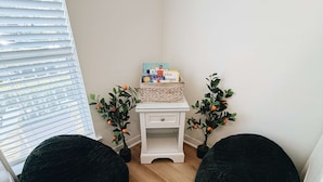 Une pièce d’angle avec une commode blanche, deux fauteuils en forme de poire noirs, une petite table blanche avec des livres et deux plantes en pot portant des fruits oranges.