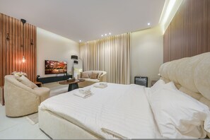 A modern hotel room with a large bed, a seating area with a sofa and armchair, a flat-screen TV, and a small table with a lamp.